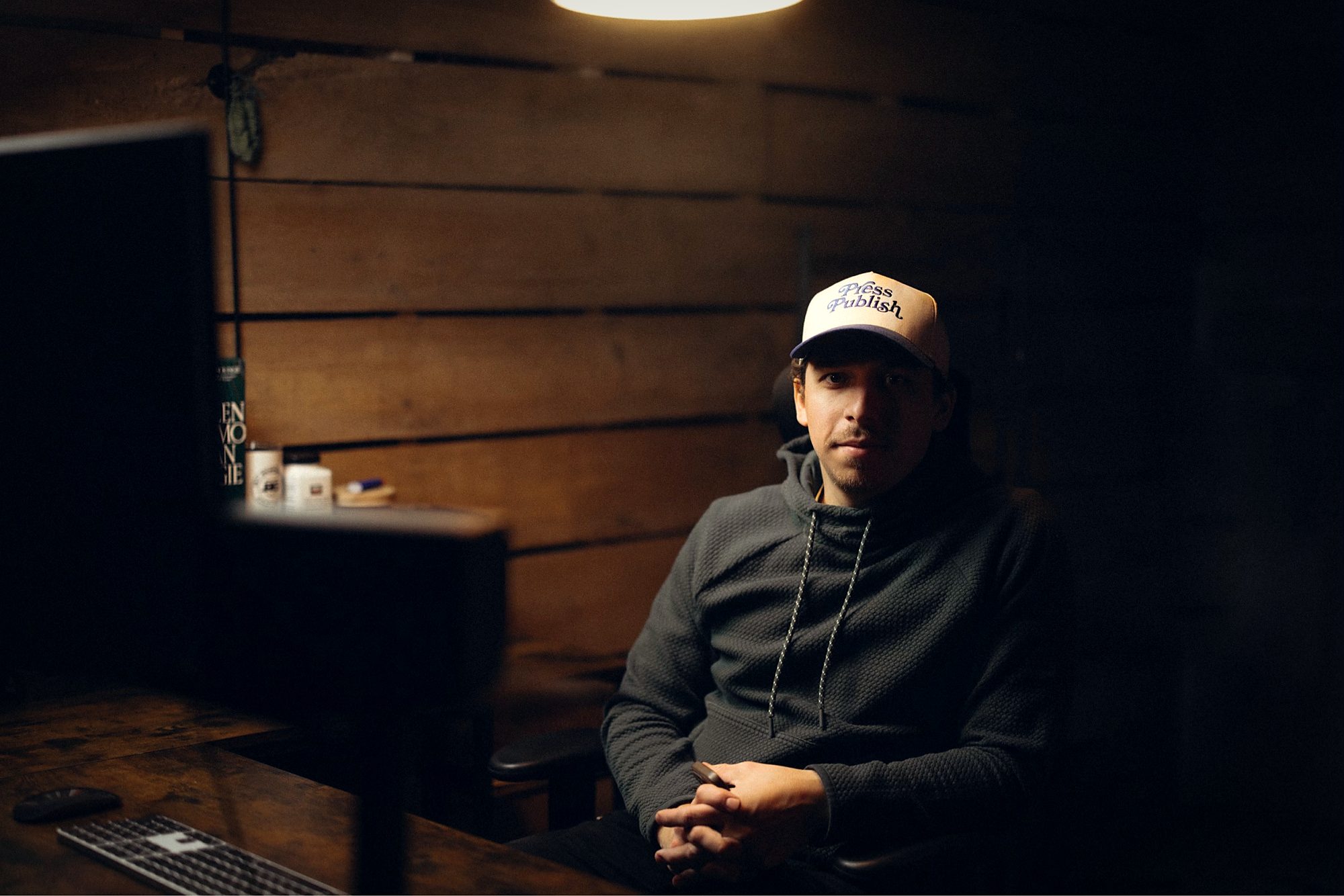 A portrait of Alek Olson at the workspace — wood-paneled studio, single overhead lamp, monitor and keyboard on the desk. Cinematic warm light, calm and considered.